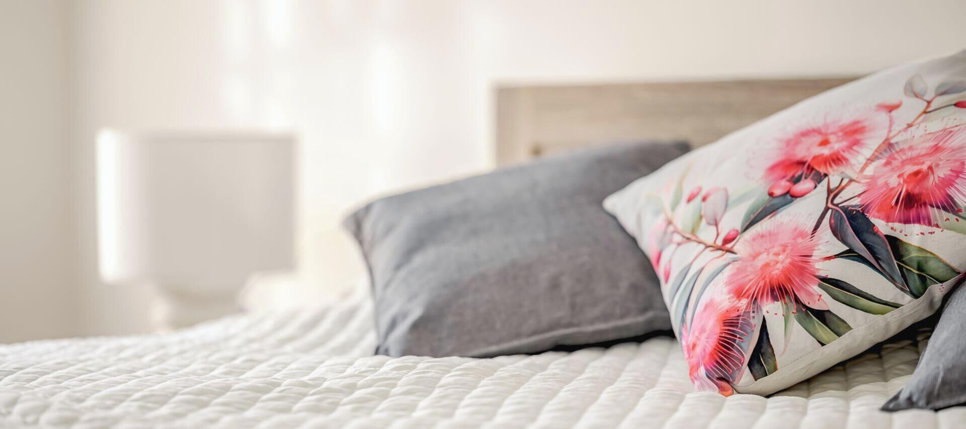 Un lit recouvert d’une couette blanche matelassée et agrémenté de deux oreillers, l’un gris et l’autre à motifs floraux.
