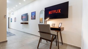 A modern hotel lobby with a flat-screen TV displaying 'NETFLIX,' a desk with a chair, and framed pictures on the wall.