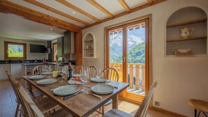 Ein Essbereich mit einem Holztisch, gedeckt für vier Personen, einer Küche im Hintergrund und Blick auf die Berge durch eine Glastür.
