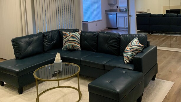 Ein Wohnzimmer mit Ecksofa, Couchtisch und einer Küche im Hintergrund.