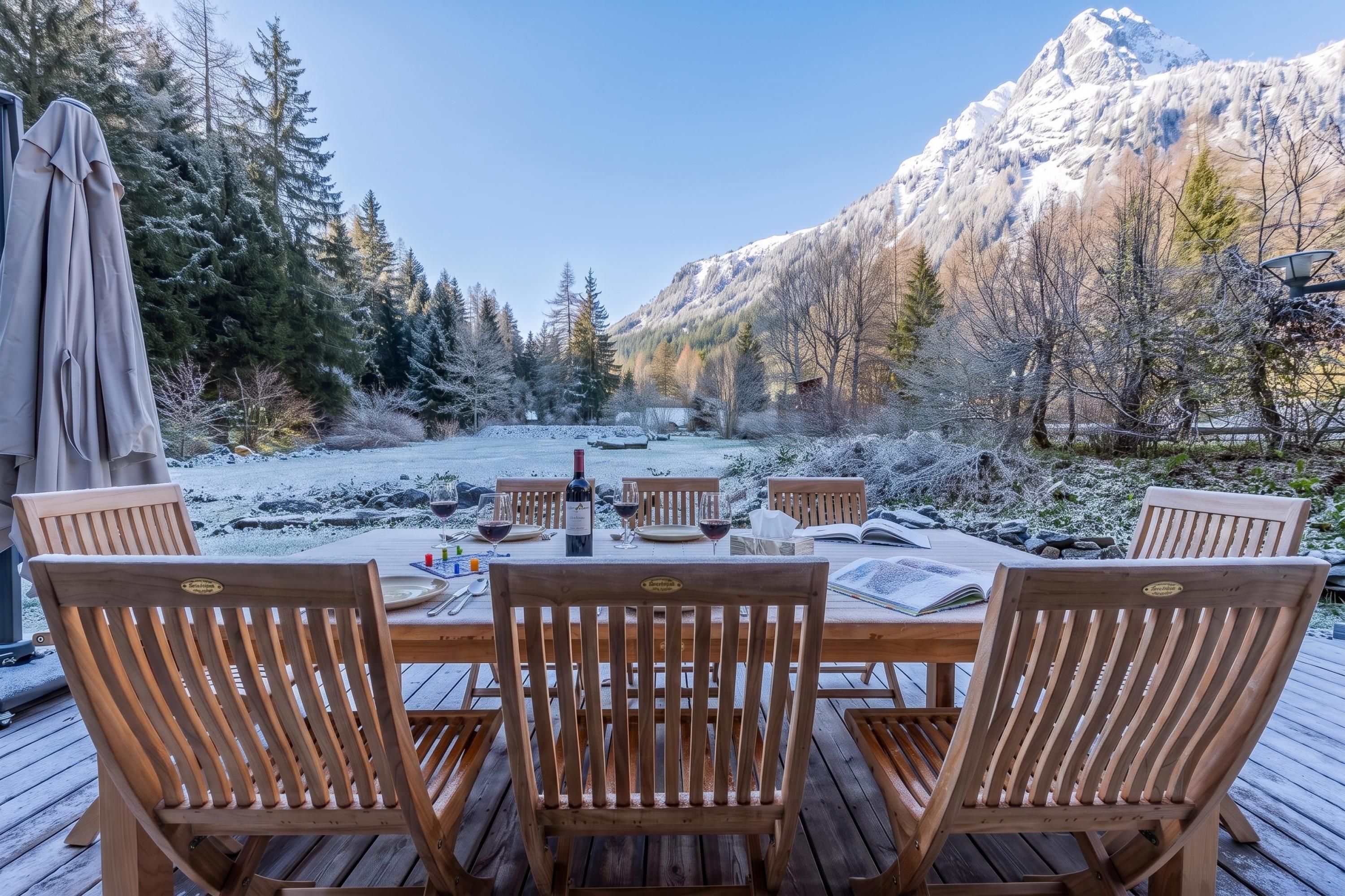 A wooden deck with a table set for dining, surrounded by chairs, overlooking a snowy landscape with trees and mountains.