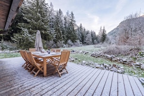 An outdoor wooden deck with a table and chairs, surrounded by snow-covered trees and a mountainous backdrop. - Chalet Montet Forest River Garden Lodge, Vallorcine (Argentiere), France (Vallorcine (Argentiere))