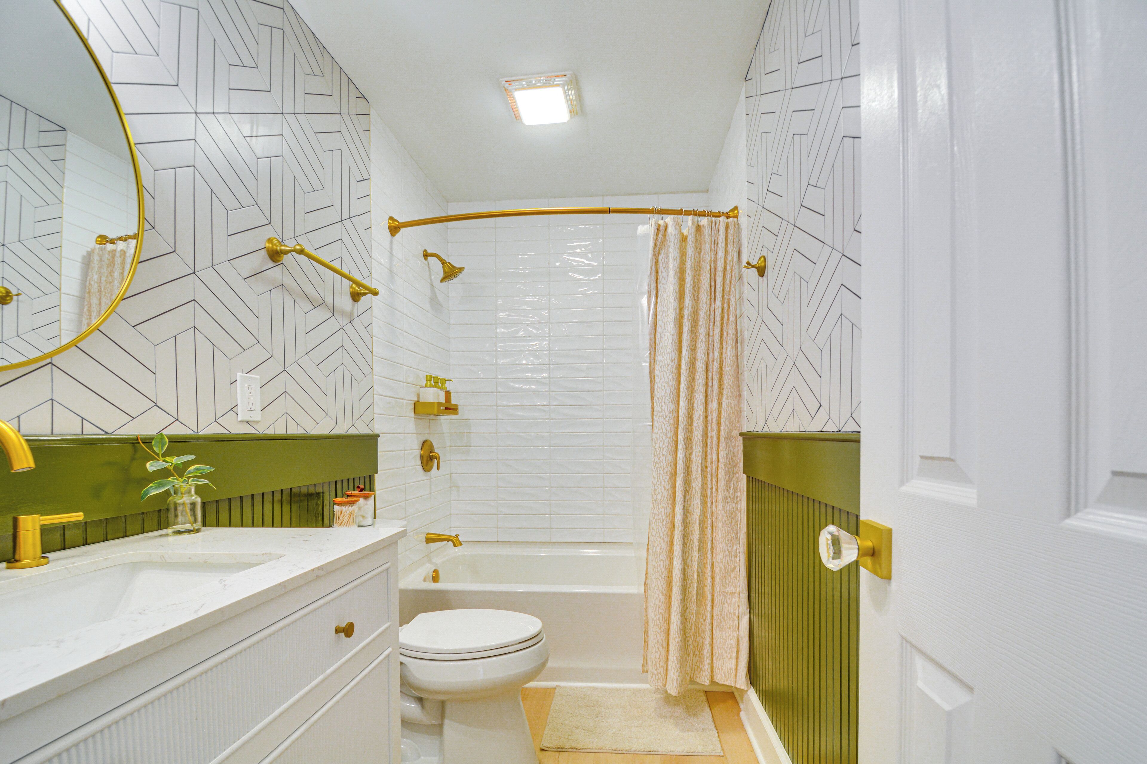 Un baño con lavamanos, inodoro y bañera blancos, con un diseño de baldosas geométricas en las paredes y cortina de ducha dorada.
