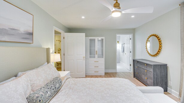 Une chambre à coucher comprenant un lit, une commode, un ventilateur de plafond et une porte donnant sur une autre pièce.