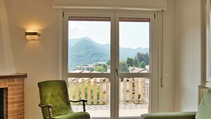 Une pièce comprenant un fauteuil à bascule recouvert d’un tissu vert, une cheminée et une porte-fenêtre coulissante donnant sur un balcon avec vue sur les montagnes et les bâtiments.