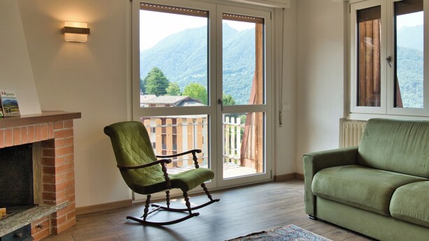 Un salon avec une cheminée, un fauteuil à bascule, un canapé vert et une vue sur les montagnes, visible à travers une porte coulissante.