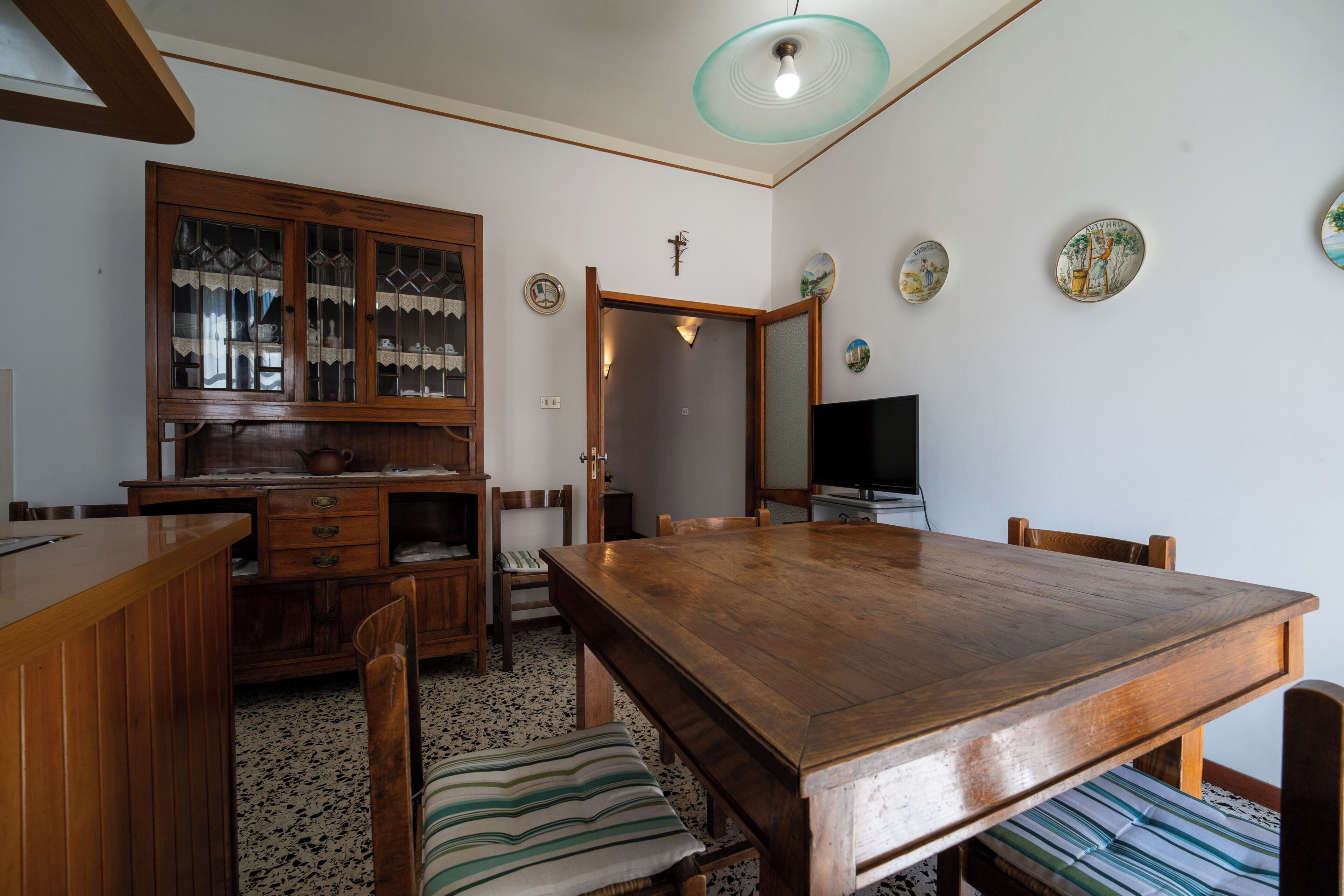 A dining area with a wooden table, chairs, a bench, a cabinet with glass doors, a television, and wall decorations.