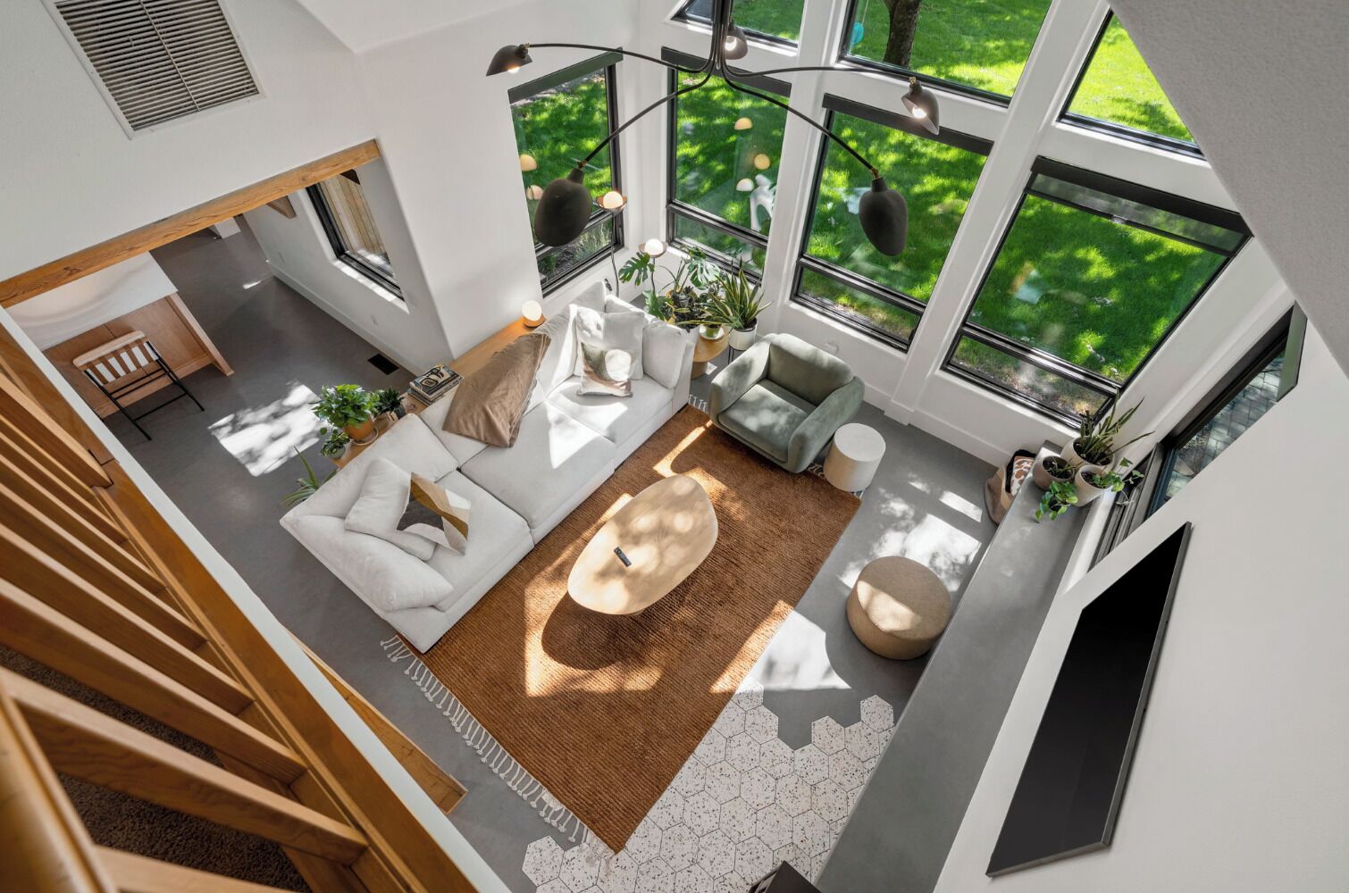 Un salon moderne avec un canapé, une table basse et des plantes, de grandes fenêtres donnant sur un paysage verdoyant, et un escalier menant à un autre niveau.