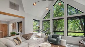 Ein modernes Wohnzimmer mit einem weißen Sofa, einem grauen Sessel, einem Couchtisch und großen Fenstern mit Blick auf eine grüne Landschaft.