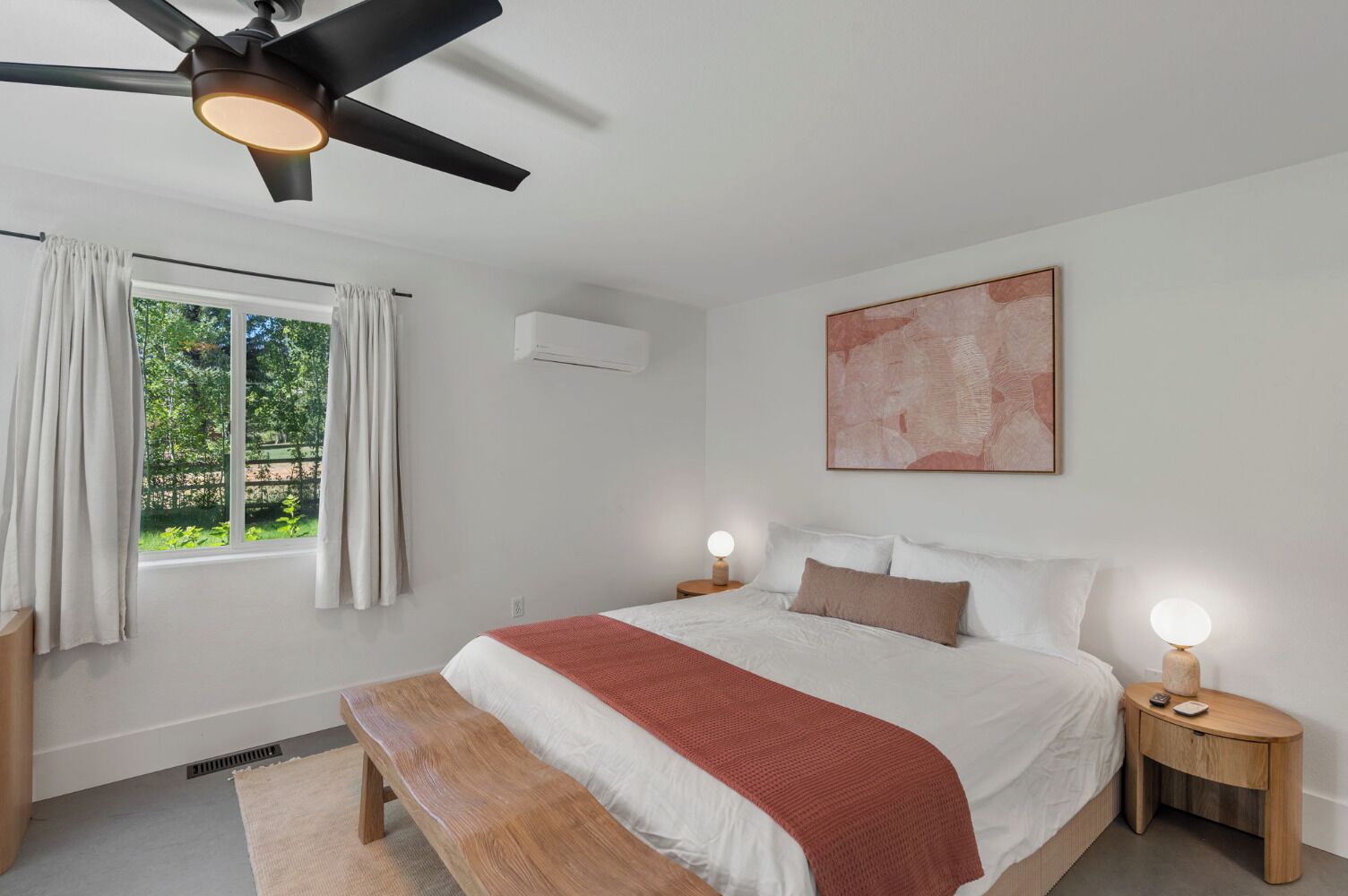 Ein Schlafzimmer mit Bett, Nachttischlampen, Deckenventilator, Fenster mit Vorhängen, Klimaanlage und einem Bild an der Wand.