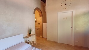 A bedroom with a bed, a bedside table with a plant, a door, and a kitchen in the background.
