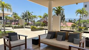 Un espacio de estar techado al aire libre con sofás y sillones, una mesita y vistas a palmeras y edificios al fondo.