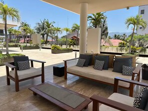 Ein überdachter Außenbereich mit Sofas und Sesseln, einem kleinen Tisch und Blick auf Palmen und Gebäude im Hintergrund.