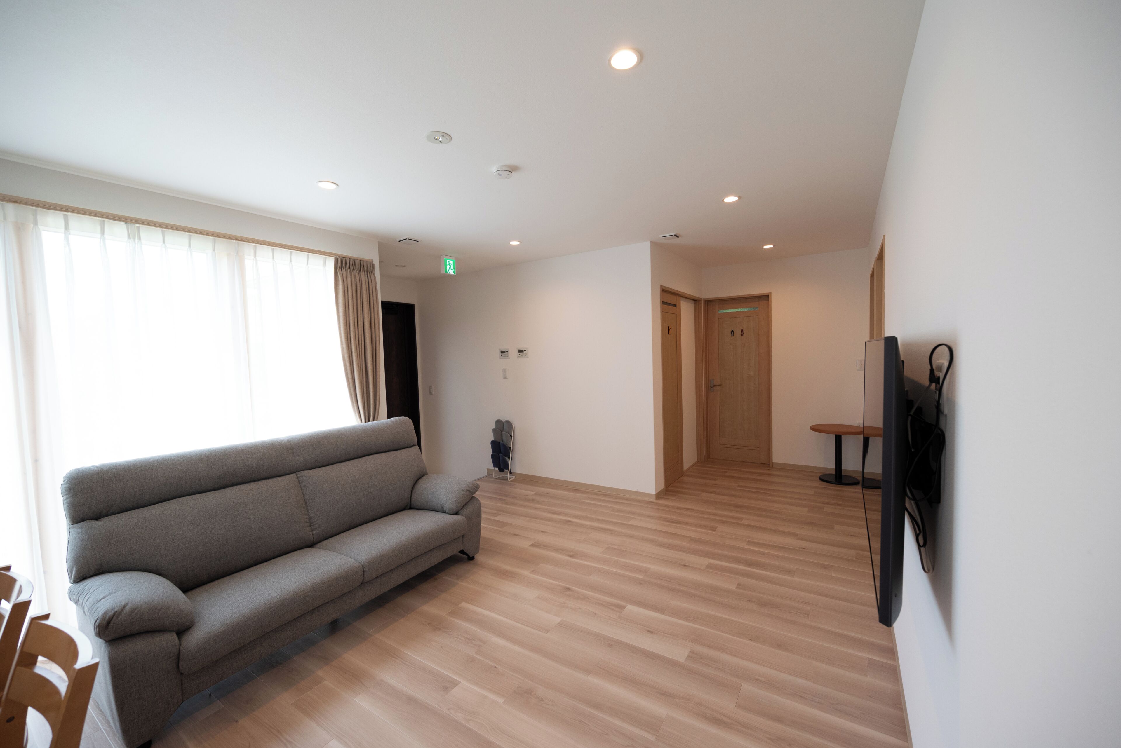 Un salón amplio con sofá, piso de madera, televisor montado en la pared y una puerta que da a otra habitación.