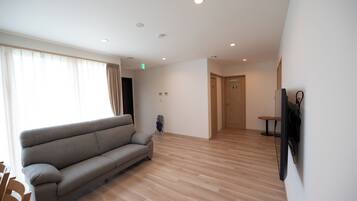 Un salón amplio con sofá, piso de madera, televisor montado en la pared y una puerta que da a otra habitación.