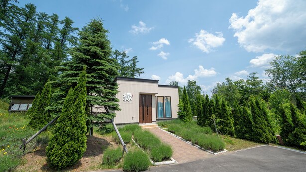 A modern building with a wooden door, surrounded by greenery and trees. - Fuyu no Sonata 2 (Nakafurano)