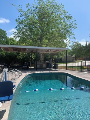 A swimming pool with clear water, a covered seating area, and a barbecue setup.