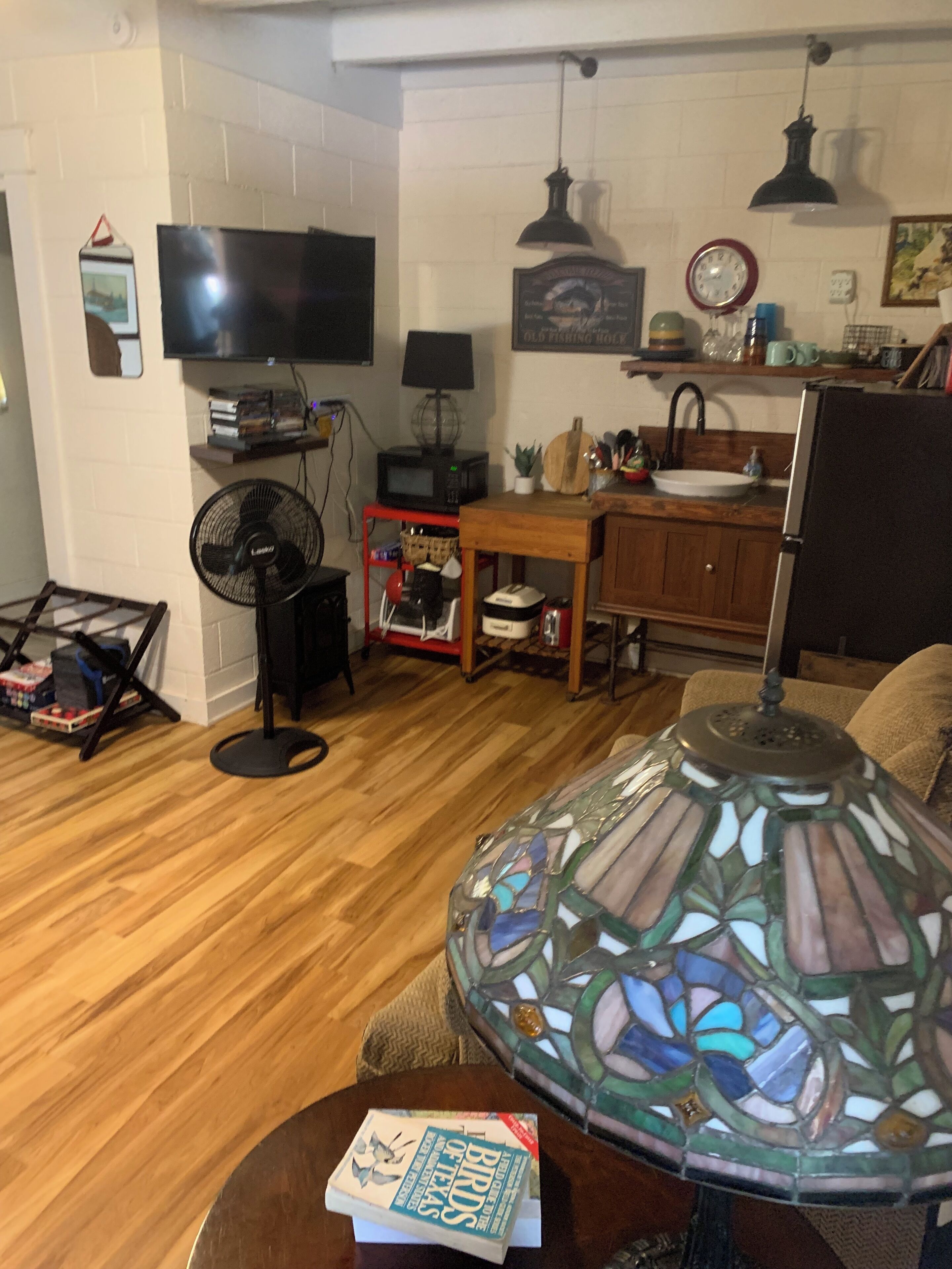 Ein Wohnzimmer mit Holzboden, einem an der Wand befestigten Fernseher, einem Tisch mit einem Buch und einer dekorativen Lampenschirm aus Buntglas.