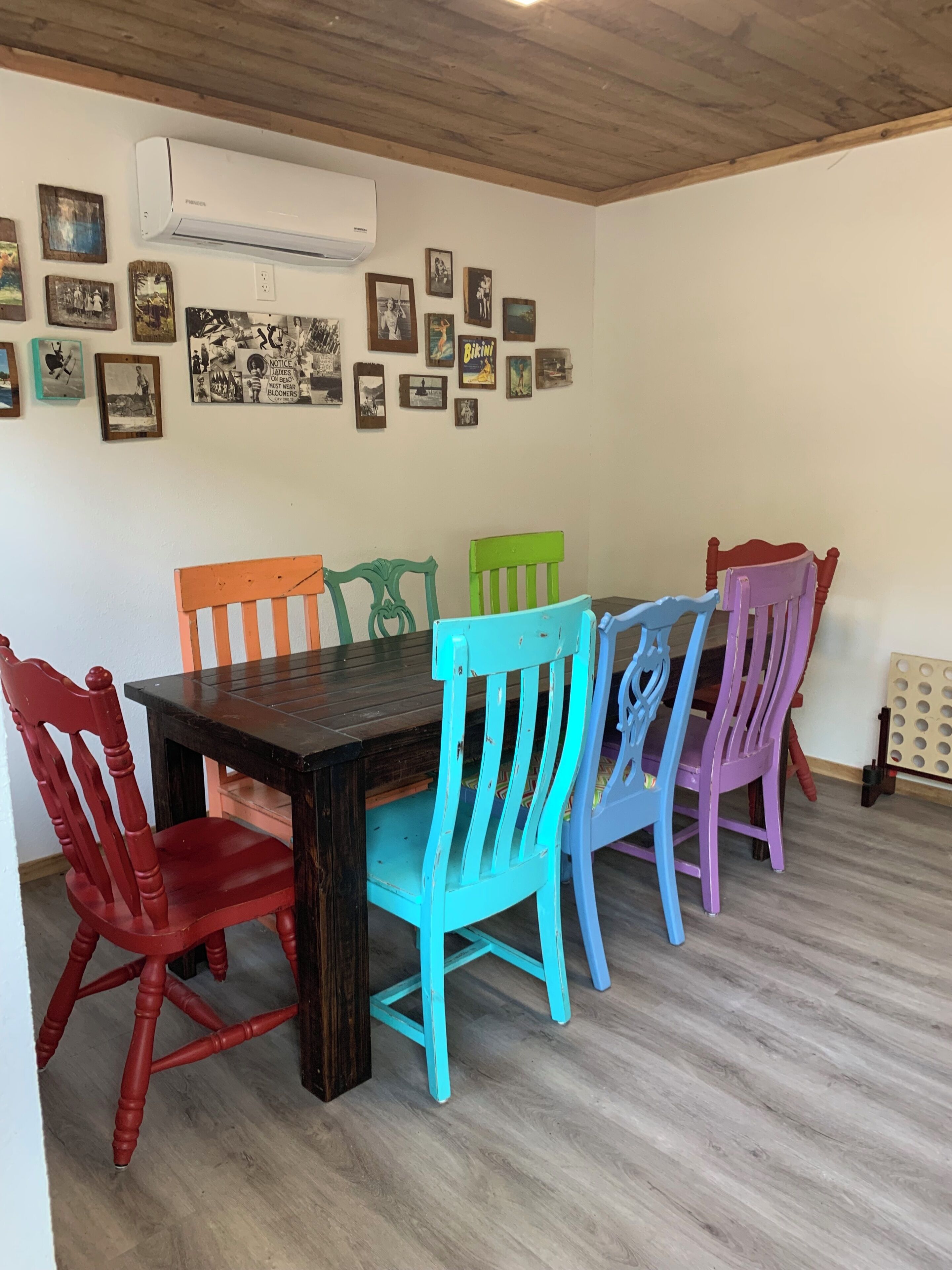 Un coin repas avec une table en bois et des chaises colorées, un climatiseur fixé au mur, et divers tableaux encadrés.
