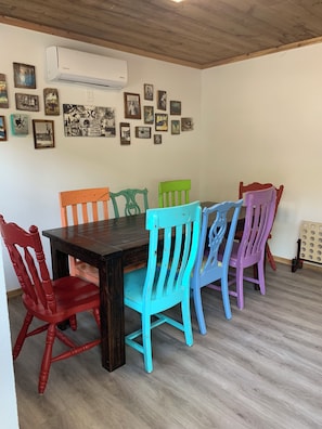 A dining area with a wooden table and colorful chairs, a wall-mounted air conditioner, and various framed pictures.