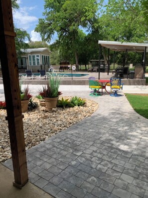 A paved patio area with potted plants, a covered seating area, and a pool in the background.