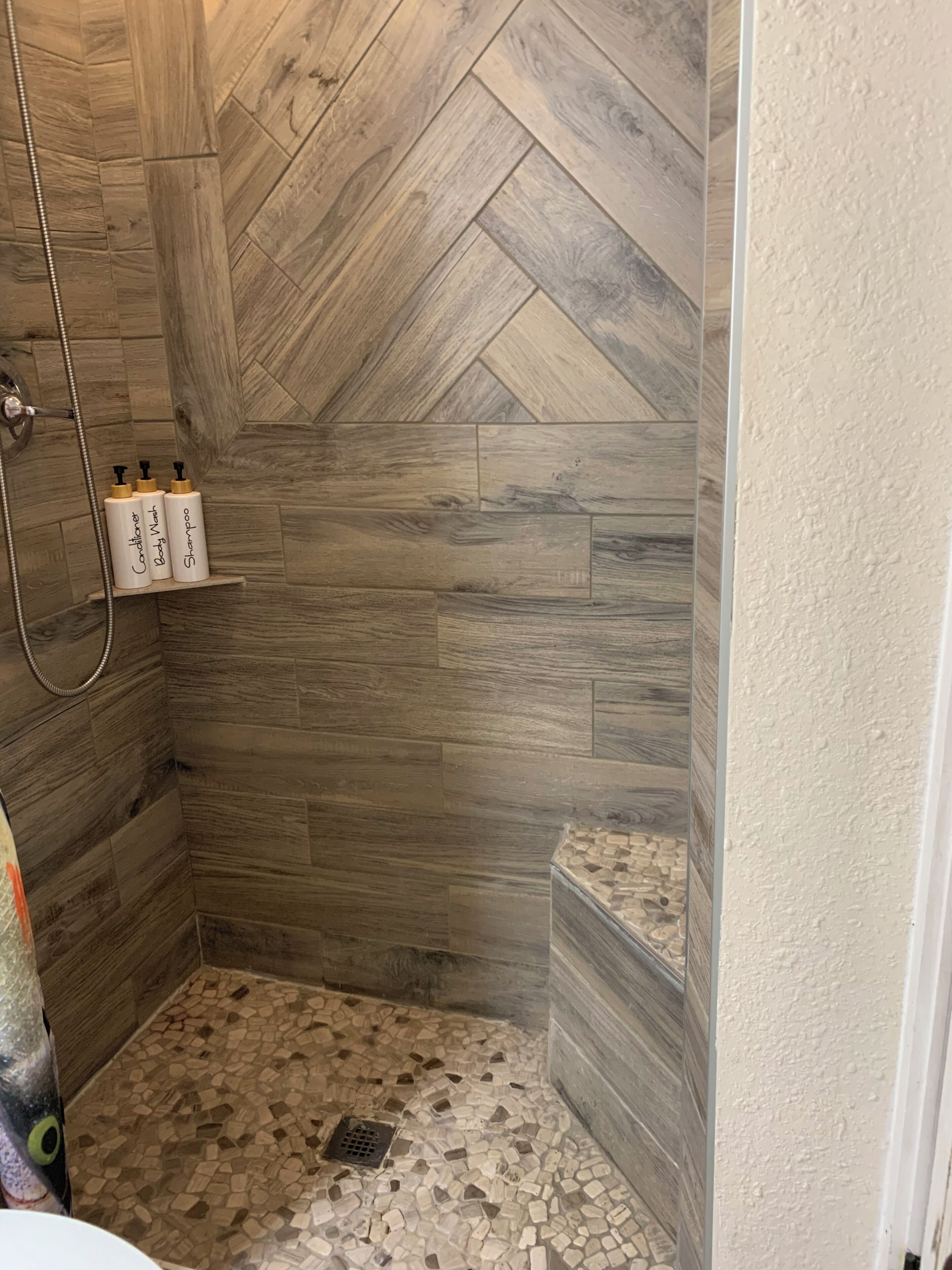 Un espace douche doté d’un sol en bois disposé en motif « point de Hongrie », d’une pomme de douche et de trois bouteilles de shampoing et d’après-shampoing posées sur une étagère.