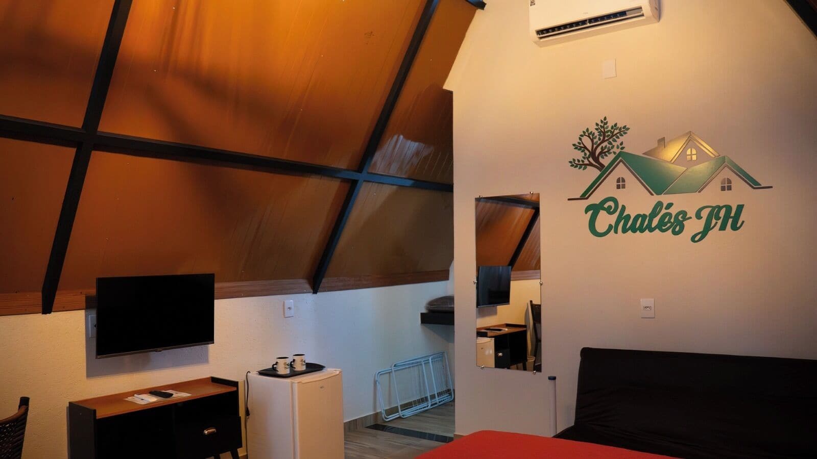 A room with a sloped ceiling, a flat-screen TV, a small refrigerator, a bed, and a wall-mounted mirror.
