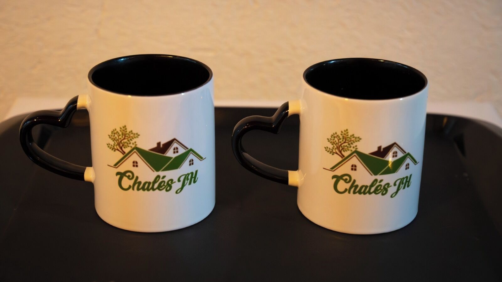 Dos tazas blancas con asas negras y diseños de casas verdes, identificadas como 'Chalès-JH'.