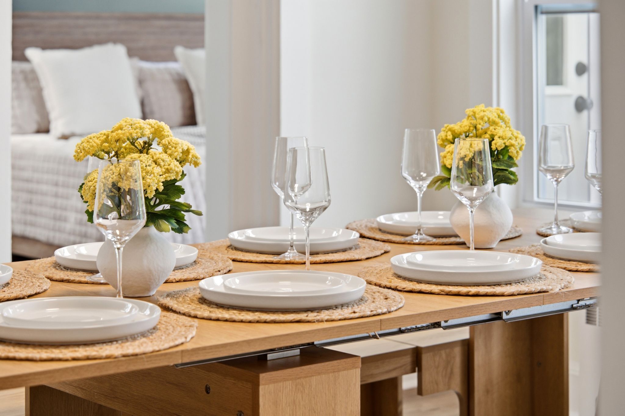 A dining table set with white plates, clear wine glasses, and yellow flowers in vases.