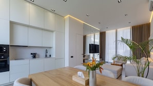 A modern kitchen with white cabinets, a wooden dining table, and a living area with a sofa and a TV. - Five Ocean Residence (Badung)