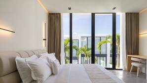A modern hotel room with a large bed, a desk, and a view of palm trees and buildings. - Five Ocean Residence (Badung)