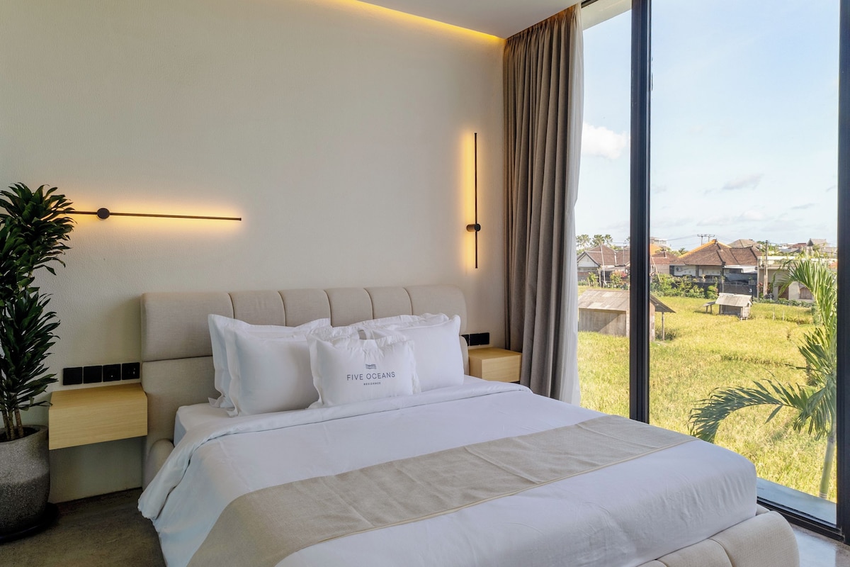 Une chambre à coucher moderne avec un grand lit, des tables de chevet, une plante en pot et une vue sur l’extérieur.