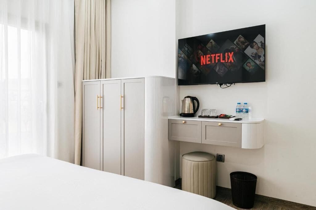 A hotel room with a bed, a TV mounted on the wall displaying the Netflix logo, a minibar, and a waste bin.