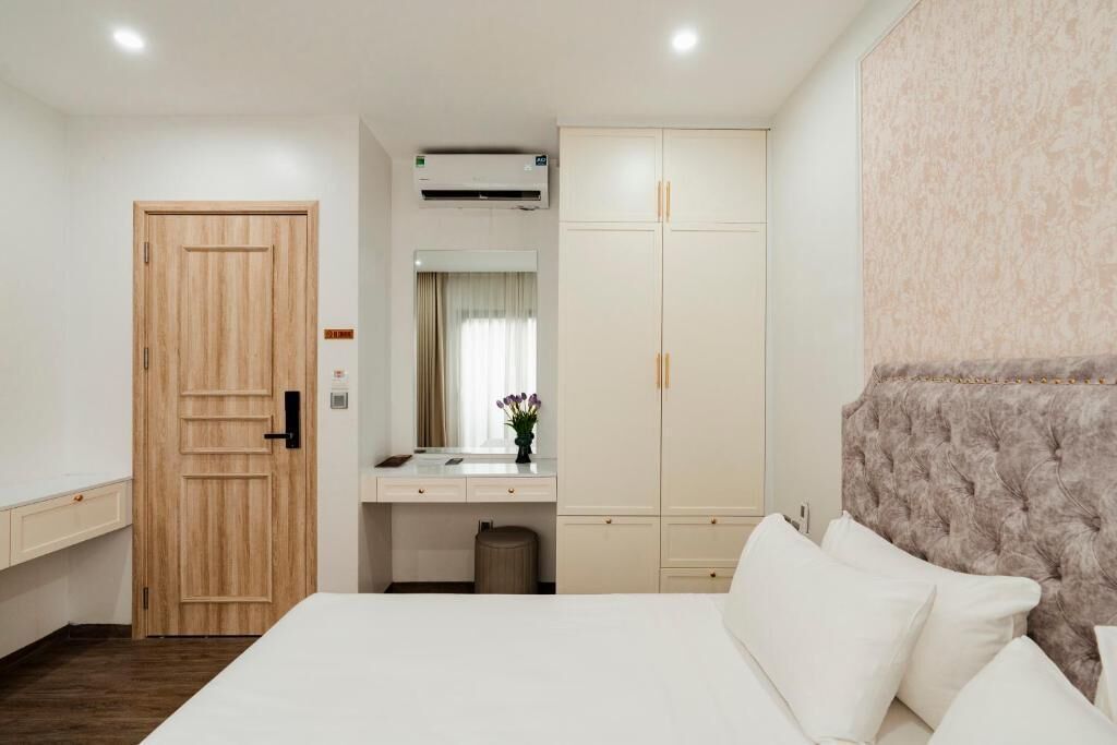 A hotel room with a wooden door, a white bed, a vanity with a mirror, a wardrobe, and a tufted headboard.