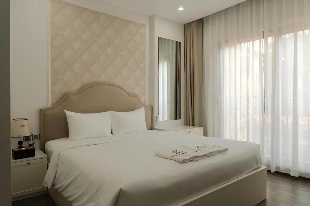 Ein Hotelzimmer mit einem großen Bett, einem Nachttisch mit Lampe, einem Spiegel und durchsichtigen Vorhängen.
