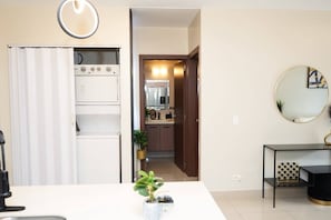 Una cocina moderna con refrigerador, horno y microondas. Hay un comedor con mesa y sillas. Un pasillo conduce a un baño con espejo y lavamanos.