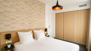 Une chambre d’hôtel avec un grand lit, une tête de lit en bois, une table de chevet avec une lampe, un luminaire fixé au mur et une armoire à portes coulissantes en bois.