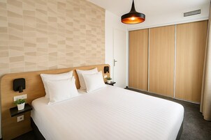 Habitación de hotel con una cama grande, cabecera de madera, mesita de noche con lámpara, luz empotrada en la pared y armario con puerta corredera de madera.