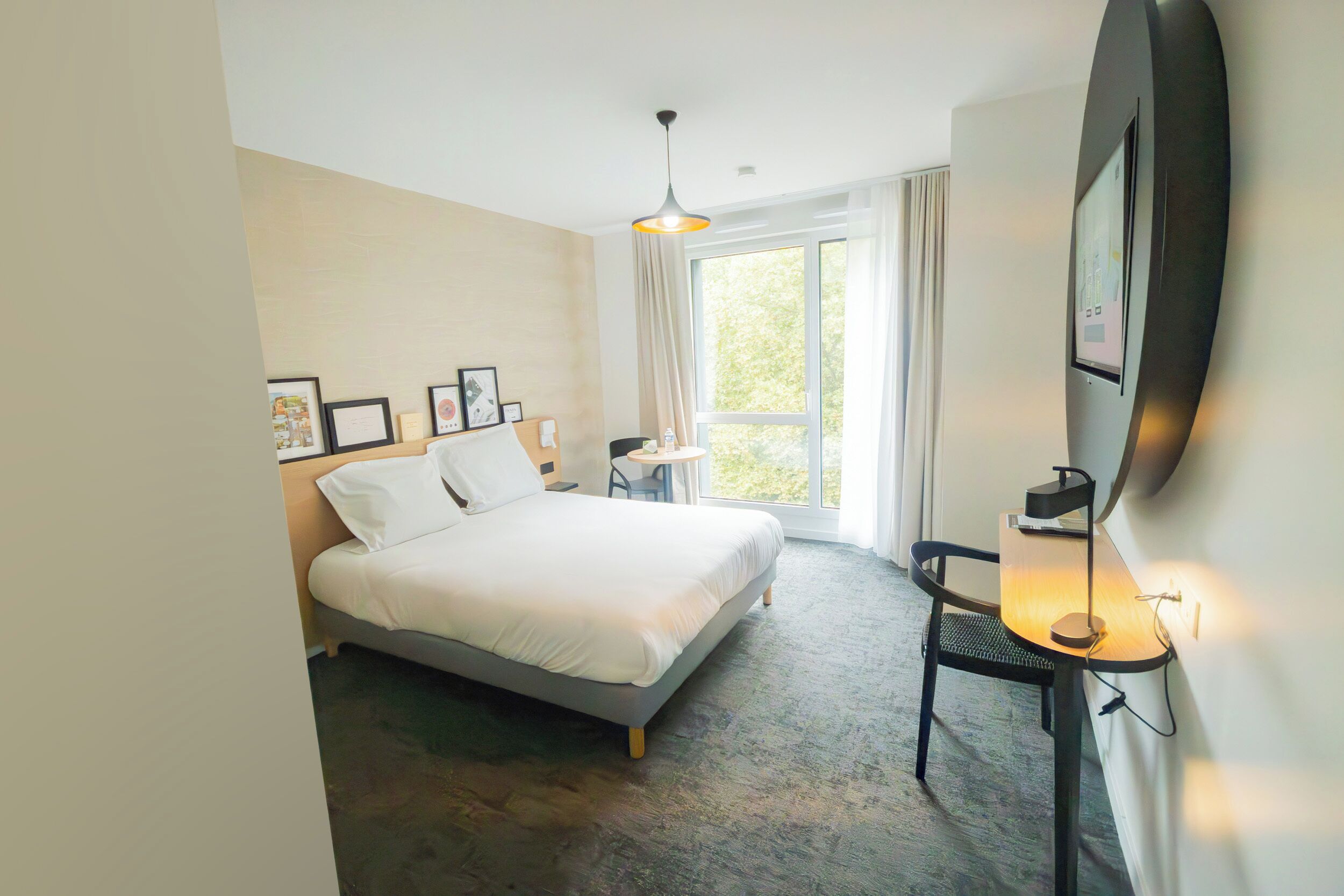 A hotel room with a bed, a desk with a lamp, a chair, a window with curtains, and framed pictures on the wall.
