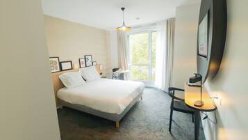 A hotel room with a bed, a desk with a lamp, a chair, a window with curtains, and framed pictures on the wall.