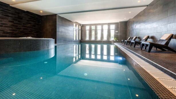 Une piscine intérieure aux murs carrelés, avec un long banc le long d’un des côtés et de grandes fenêtres laissant entrer la lumière naturelle.
