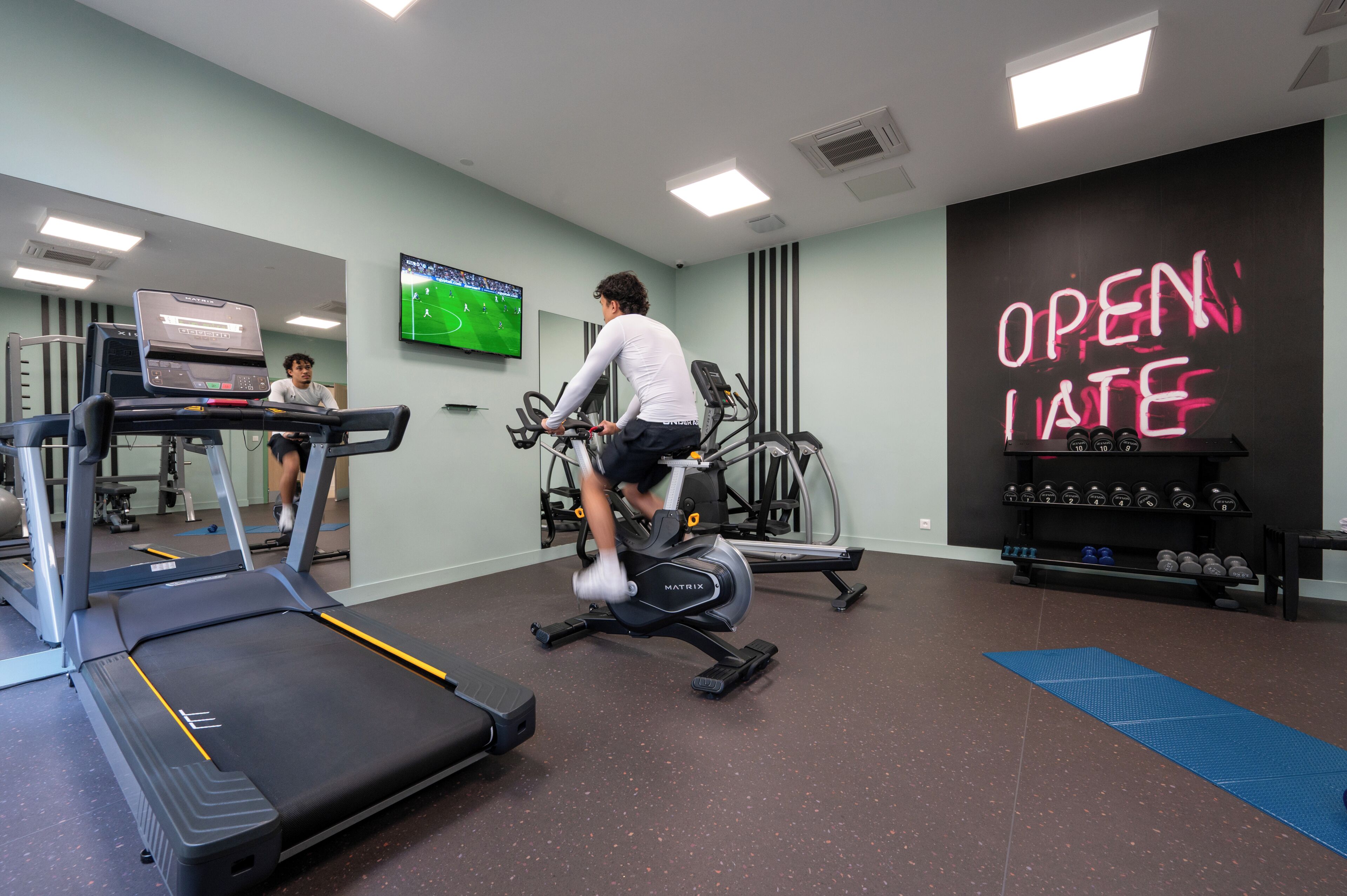 A gym with treadmills, an elliptical, and a wall-mounted TV displaying a soccer game.