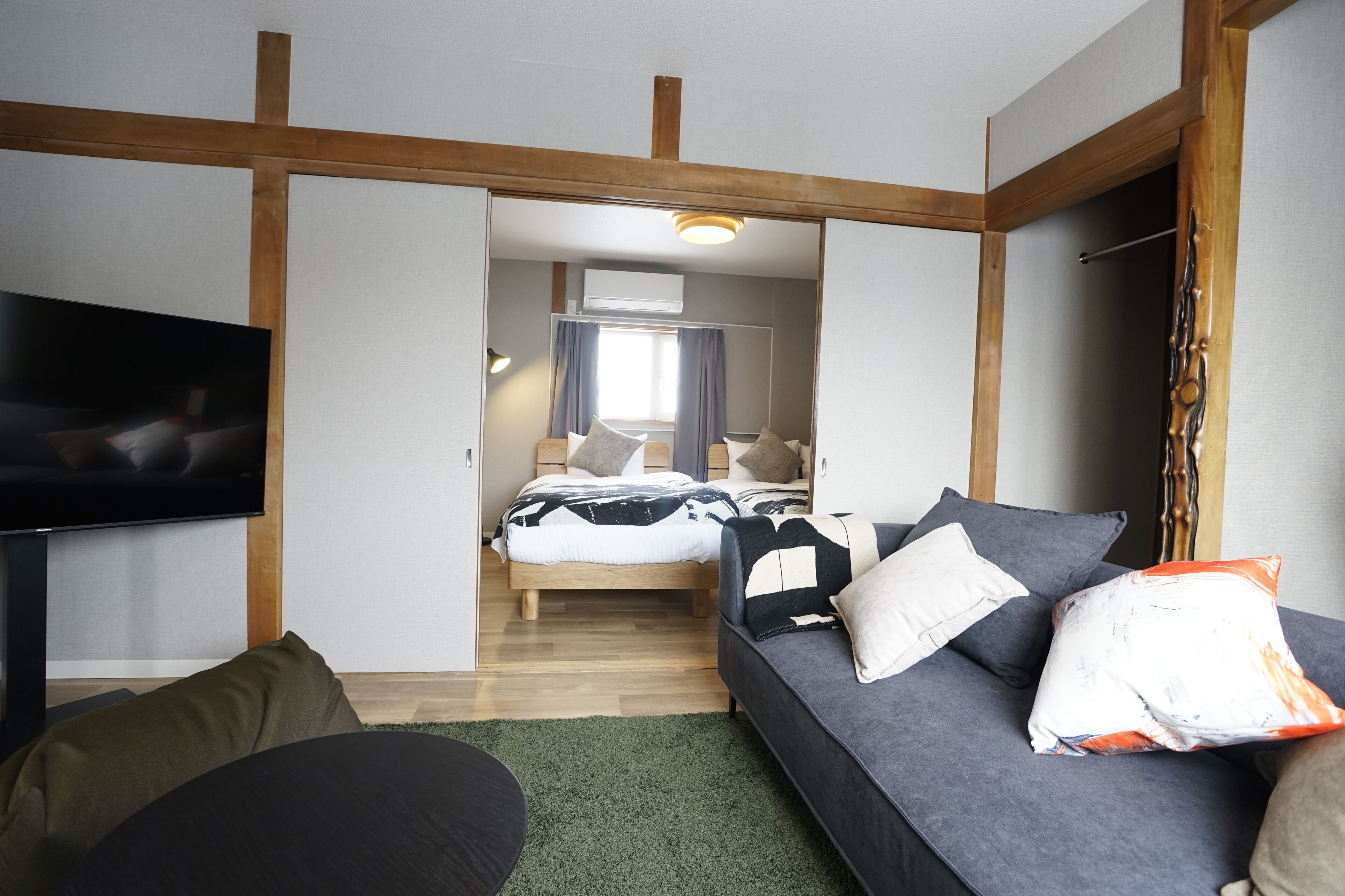 A modern hotel room with a bed, a sofa, a TV, and a wooden ceiling structure.