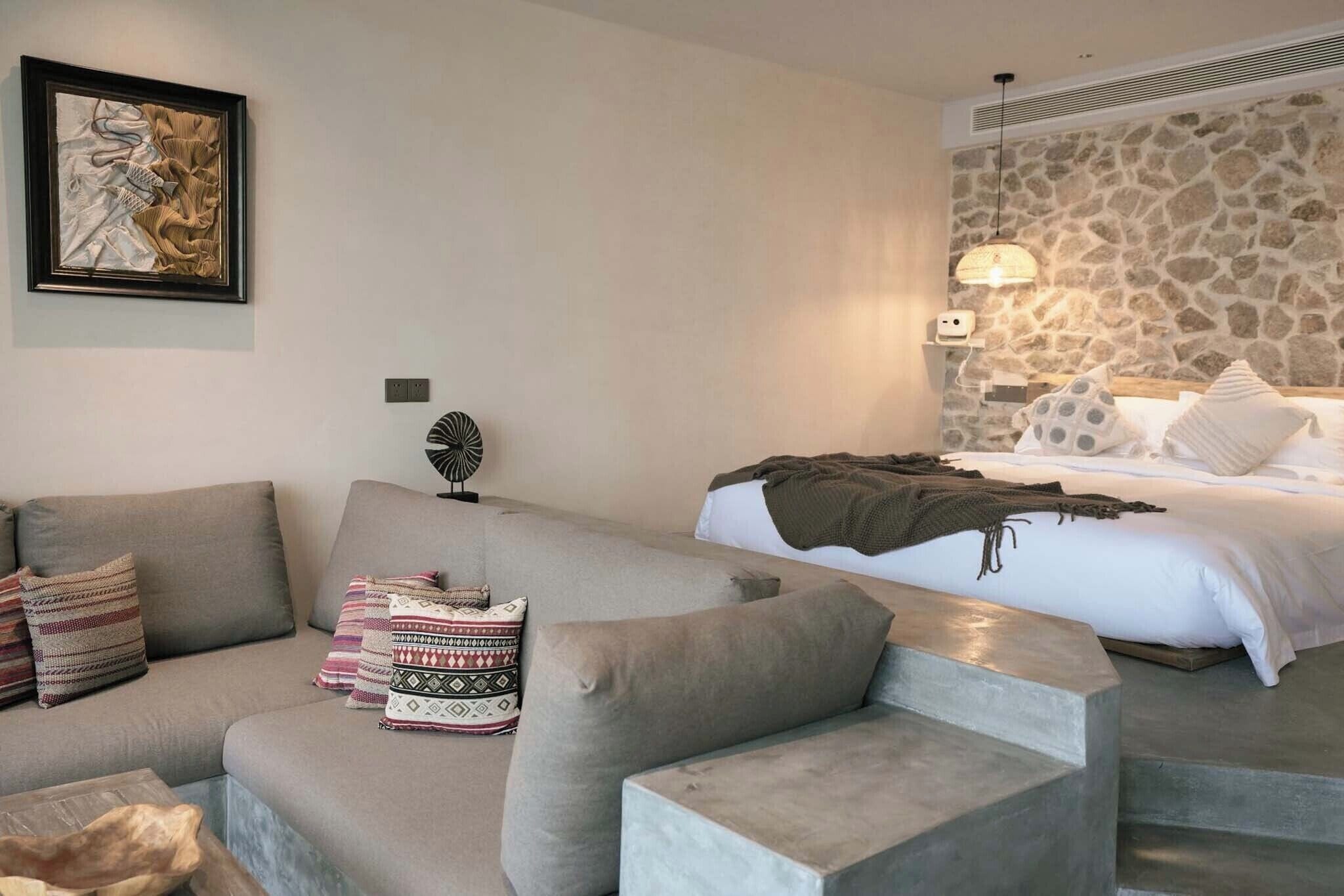Una habitación de hotel moderna con un sofá, una cama con almohadas estampadas, una pared de piedra y una obra de arte enmarcada en la pared.