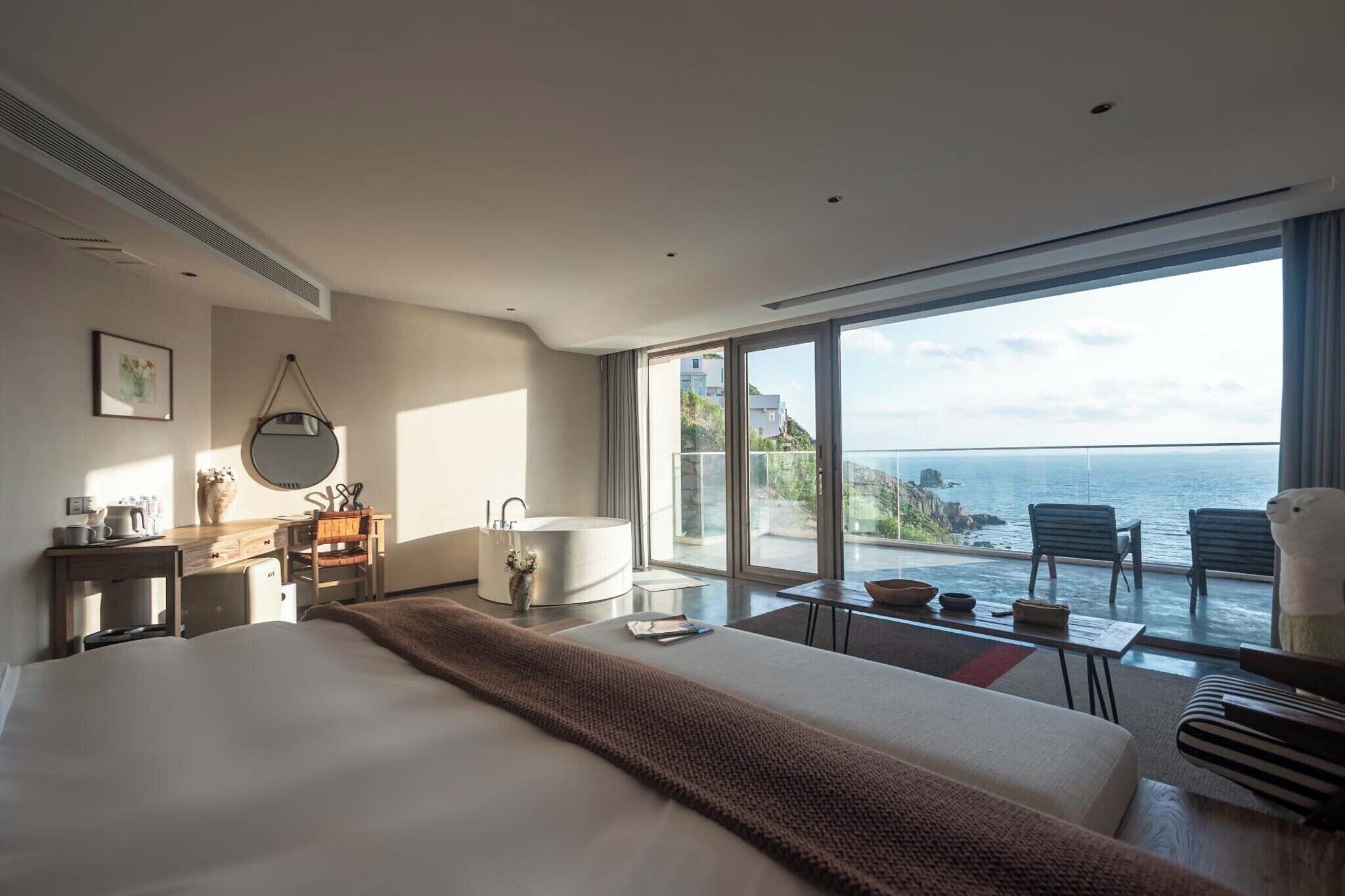 A modern hotel room with a large bed, a sitting area with a view of the sea, a bathtub, and a desk with a mirror.