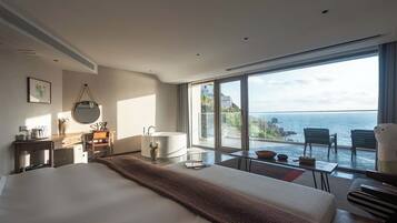 A modern hotel room with a large bed, a sitting area with a view of the sea, a bathtub, and a desk with a mirror.