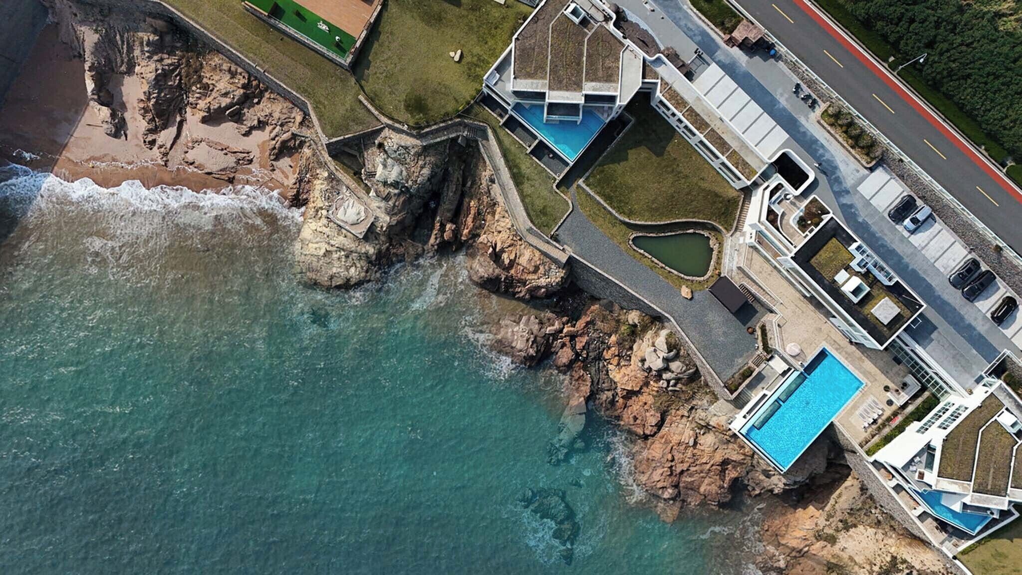 Vue aérienne d’une propriété située sur la côte, avec une piscine, une route et une plage rocheuse.