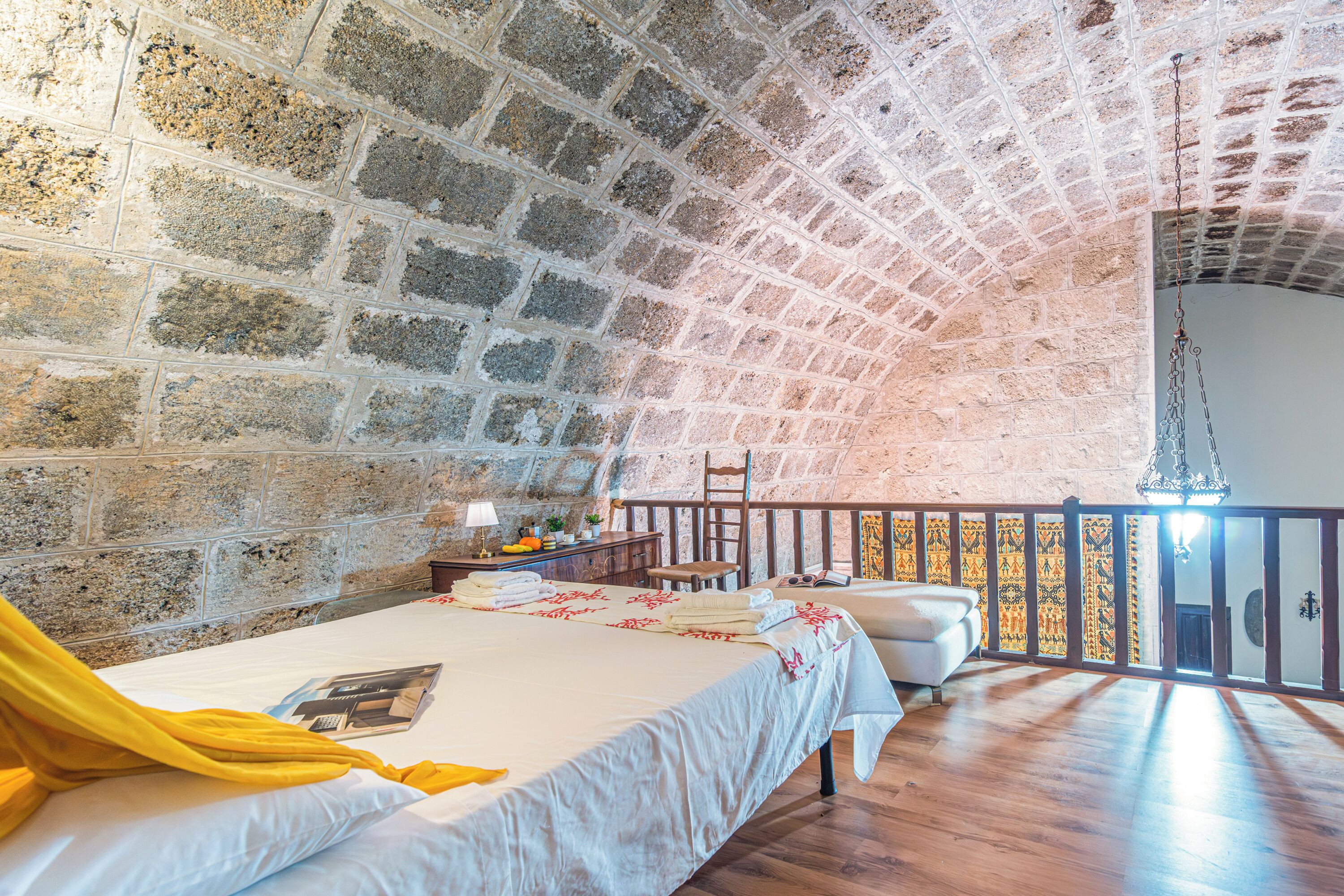 A room with a stone wall, a bed with a yellow blanket, a wooden floor, and a balcony with a railing.