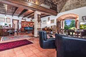 A spacious living room with a piano, leather sofas, a dining area, and a view of greenery through a large window. - Fort Galantè, Alghero, Italy (Alghero)