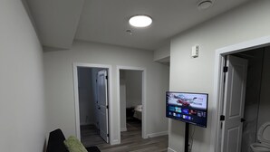 A modern living room with a black sofa, a small table, a flat-screen TV, and a hallway leading to a bedroom. - Cozy 2BR Suite Near NAIT | Wi-Fi + Kitchen (Edmonton)